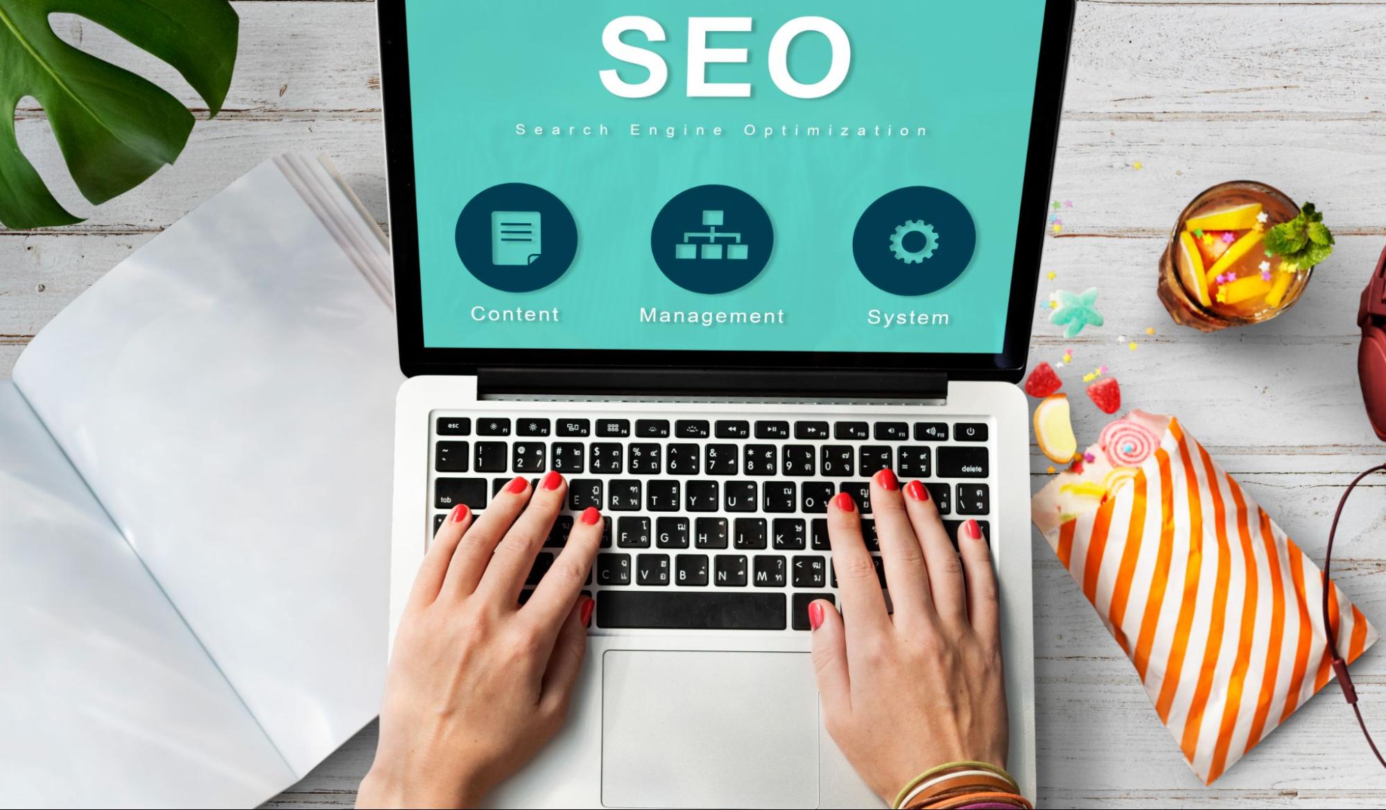 Woman with red nails working on SEO at a laptop, with coffee and juice beside her, a smart alternative to a traditional SEO agency for startups.