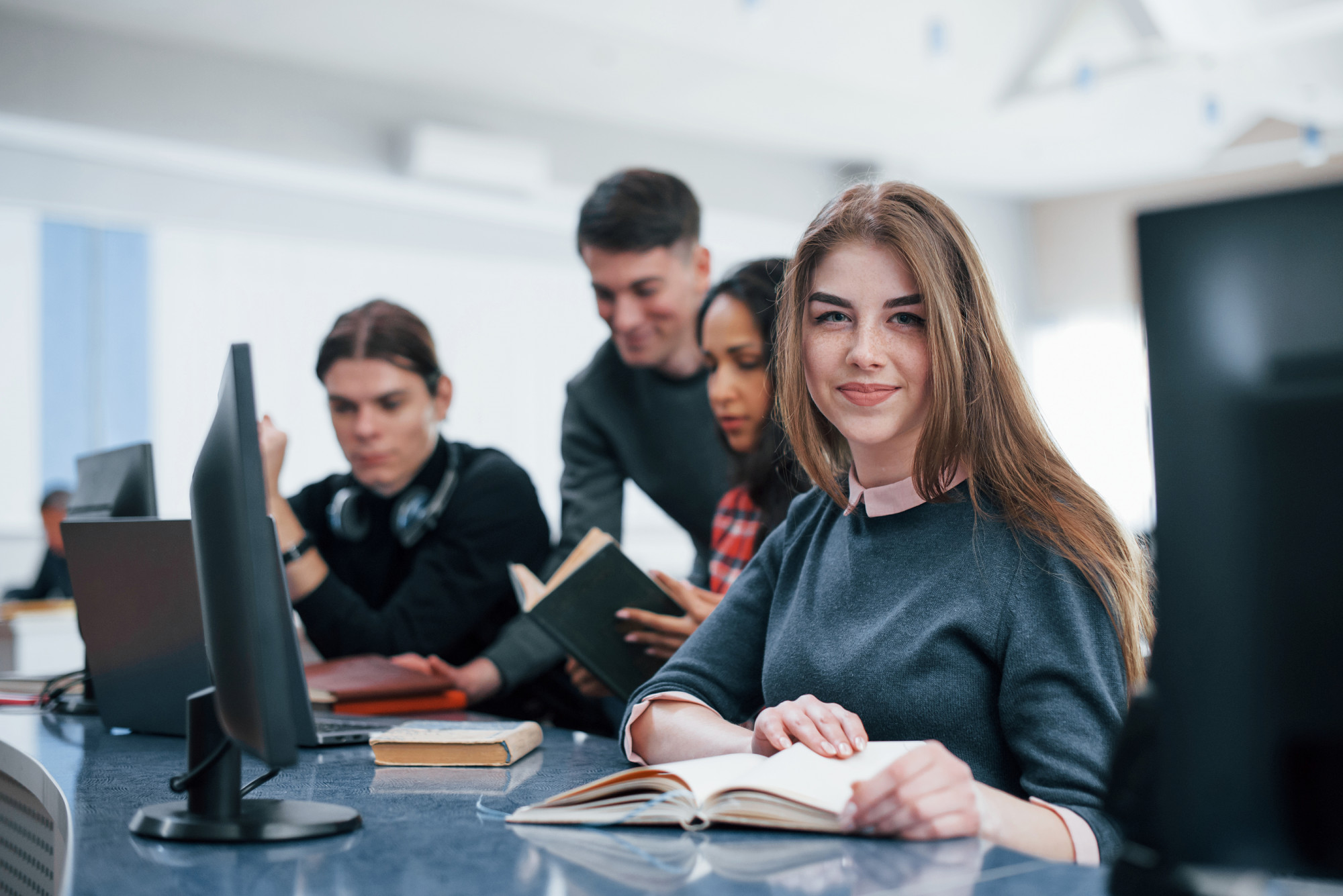 A young woman in a library smiles at the camera while reading. Three colleagues are behind her, engaged with books and a computer. Bright, focused atmosphere.