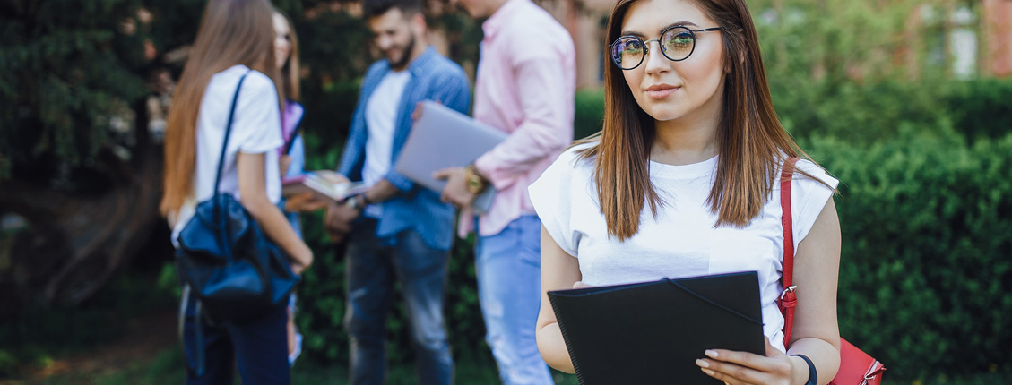 A young woman with glasses holds a folder, standing confidently in a grassy area. In the background, a group of friends chat, creating a relaxed, academic atmosphere.