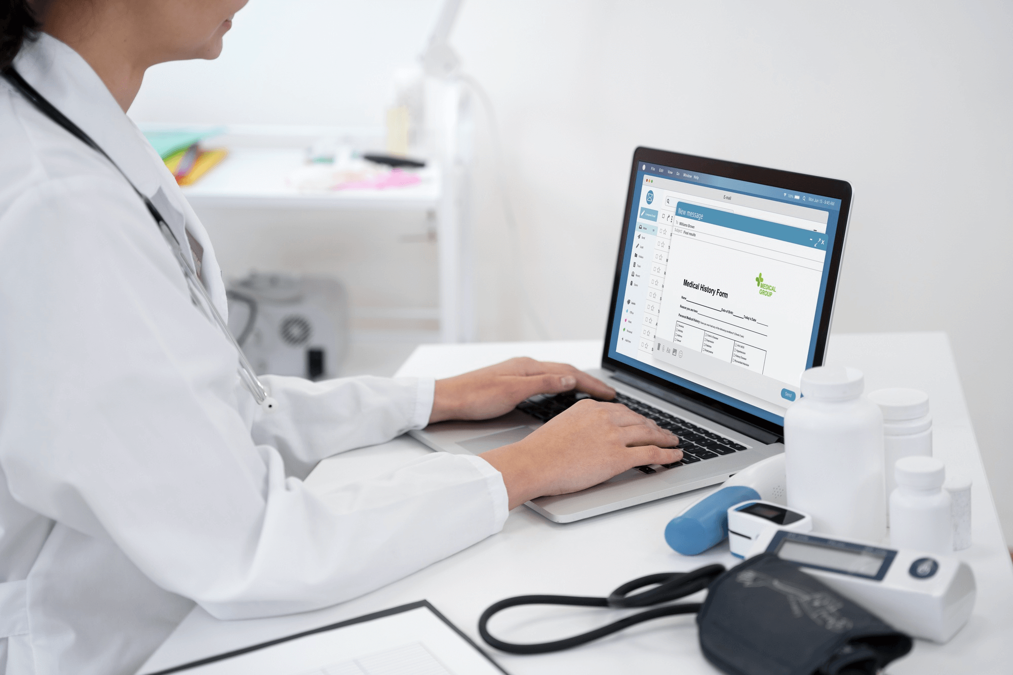 A doctor in a white coat types on a laptop displaying a medical history form. The desk holds medicine bottles, a blood pressure monitor, and medical tools.