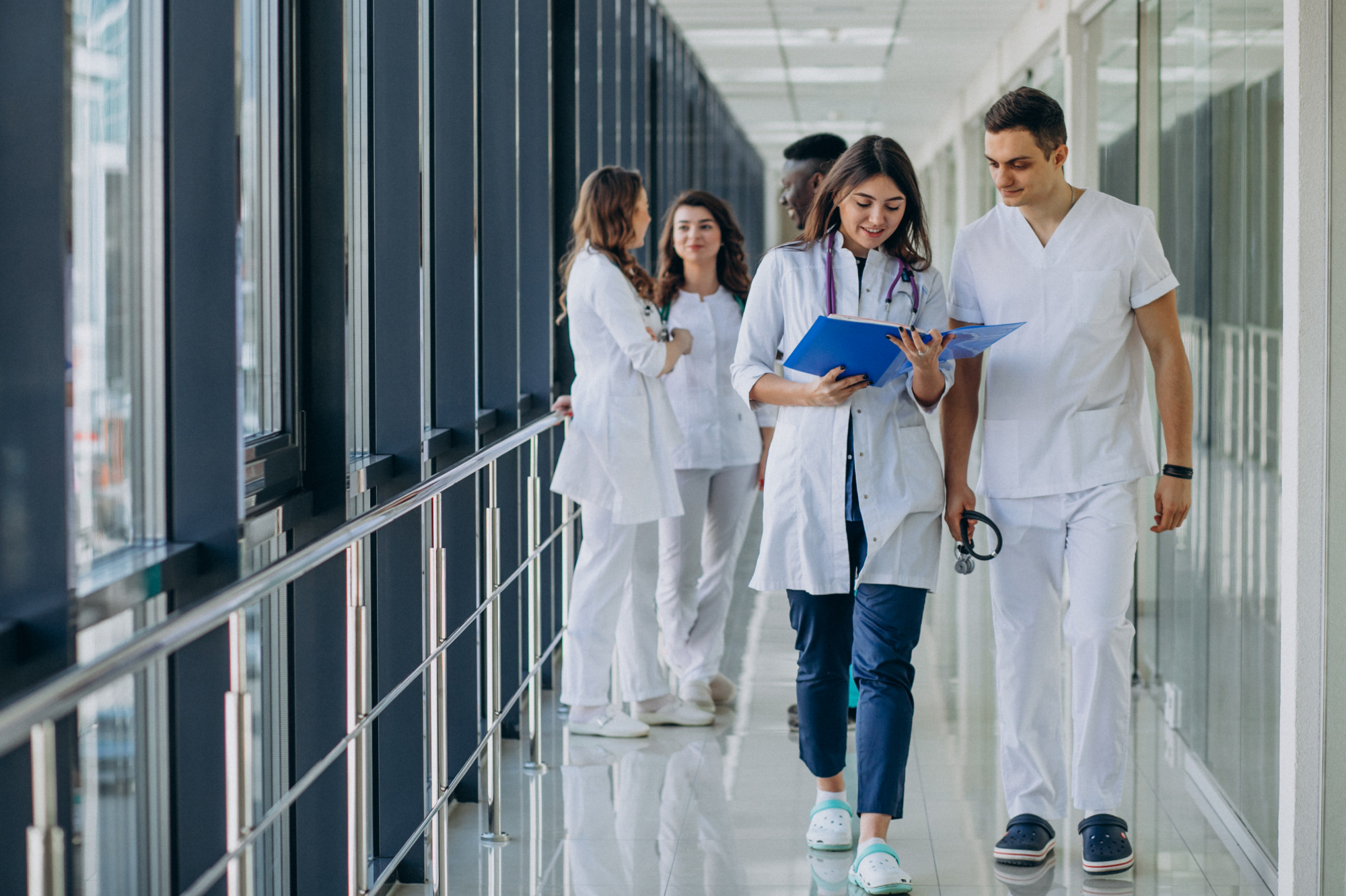 A group of healthcare professionals in white uniforms walk down a hospital corridor. Two in front discuss a blue file, conveying teamwork and focus.