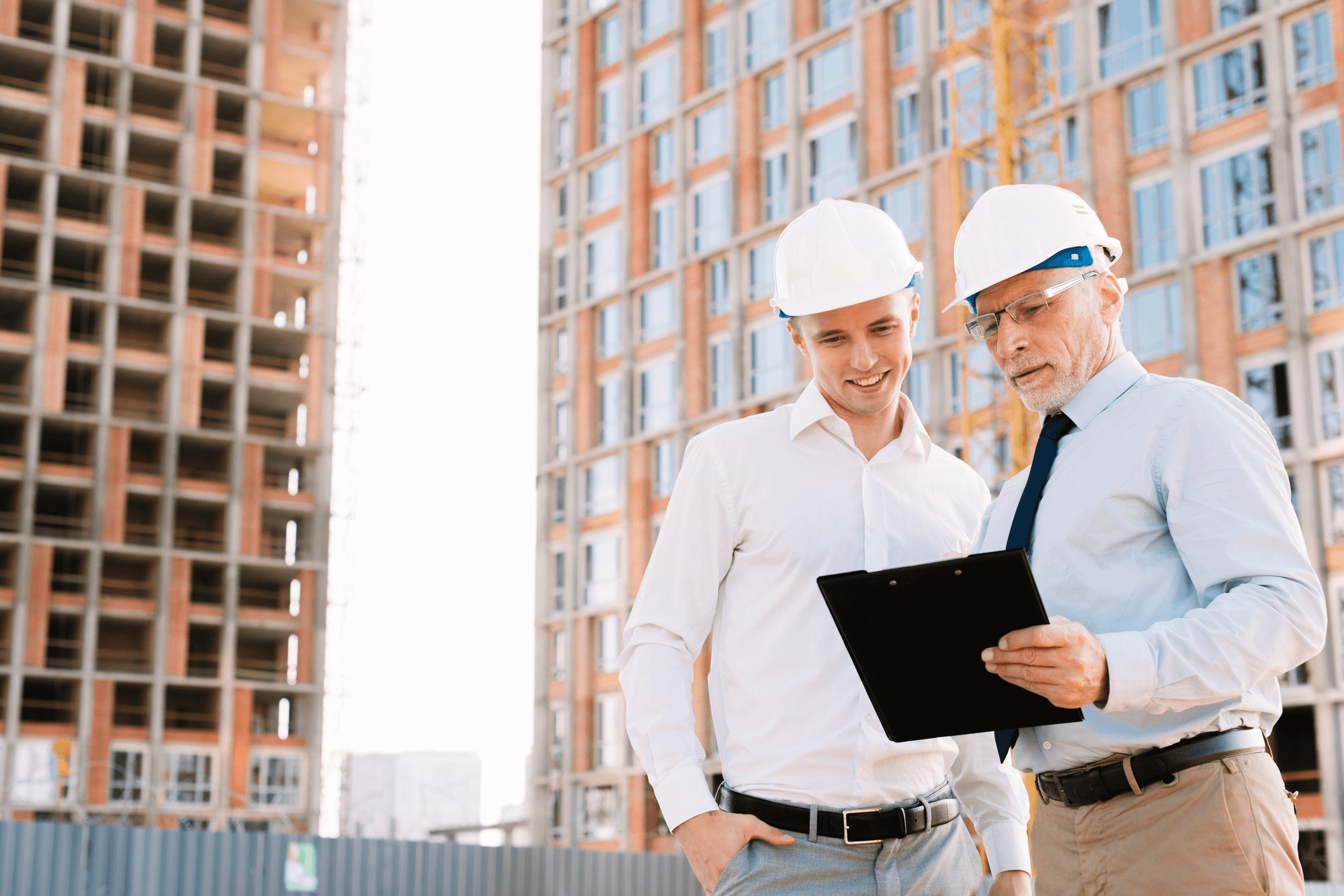Two men in white shirts and hard hats review plans on a clipboard at a construction site. A partially-built building stands in the background.