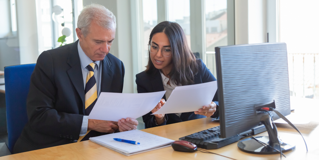 A man and woman in business attire discuss documents at a desk with a computer. They appear focused and engaged, set in a bright, modern office.