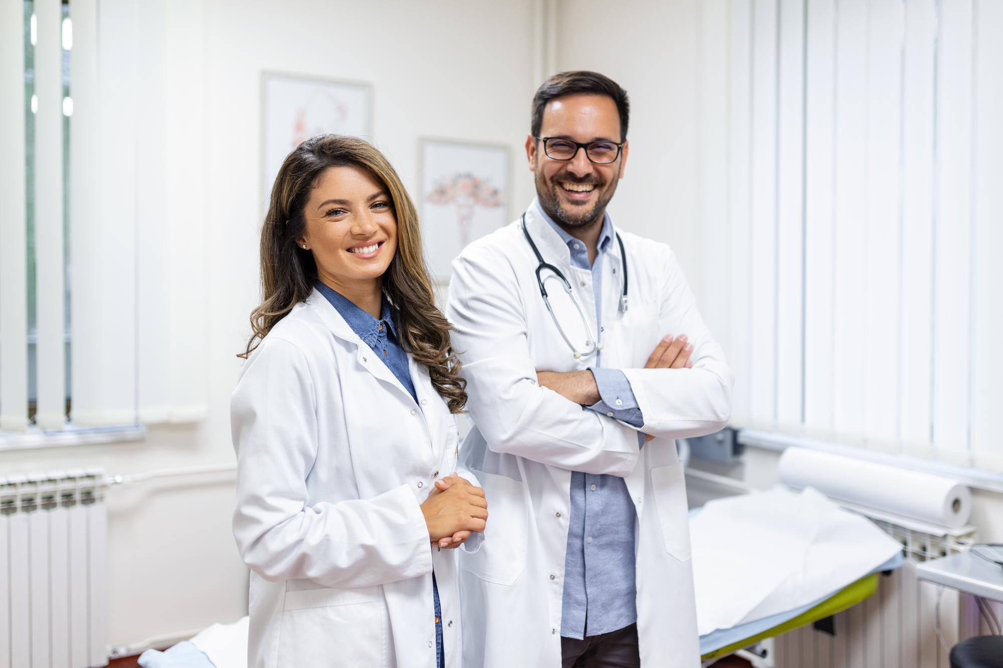 Two smiling medical professionals, a man and a woman, wearing white coats and stethoscopes, stand confidently in a bright, modern exam room.