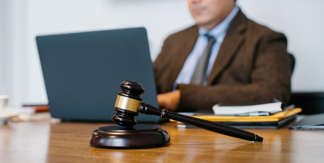 A wooden gavel rests on a desk in the foreground. In the blurred background, a person in business attire is working on a laptop, suggesting a legal setting.