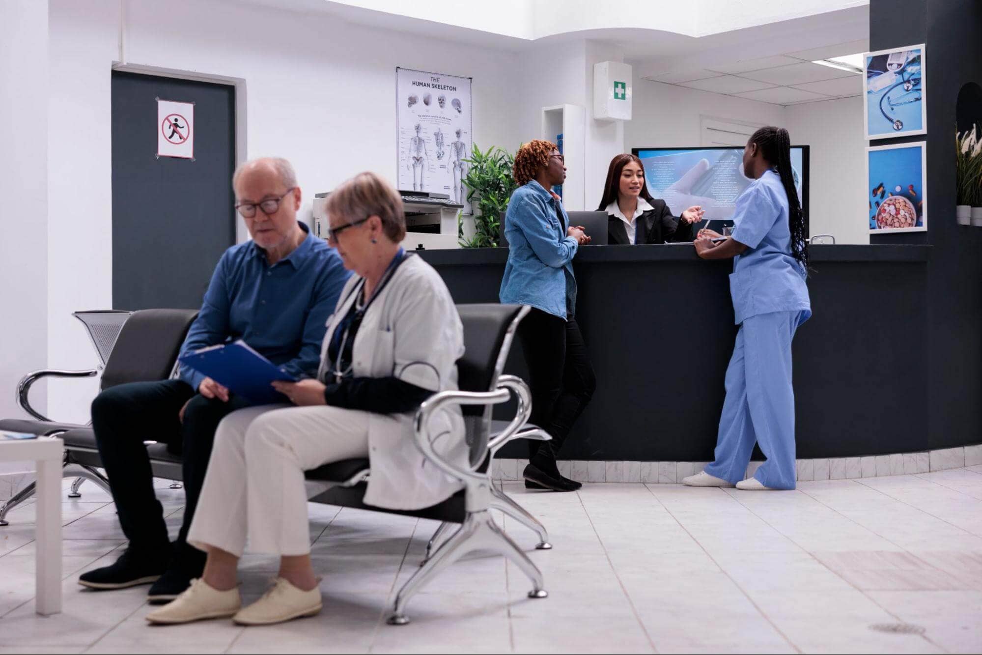 Doctor checking a wheelchair-bound patient, another talking to the receptionist, and one reading a brochure—capturing a busy clinic boosted by urgent care SEO.
