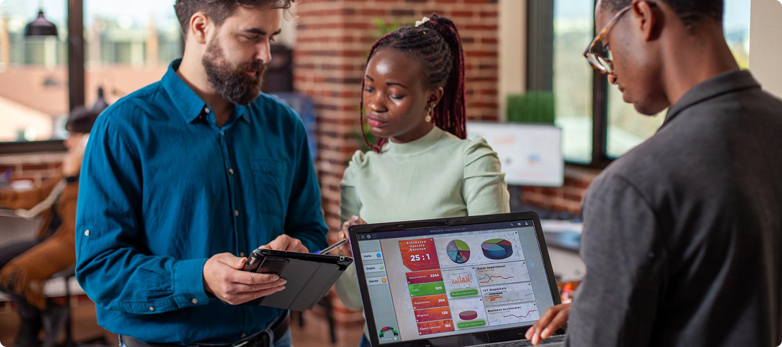 Three people in an office review data on a tablet and laptop showing charts and graphs. The setting is professional, with a focus on teamwork.